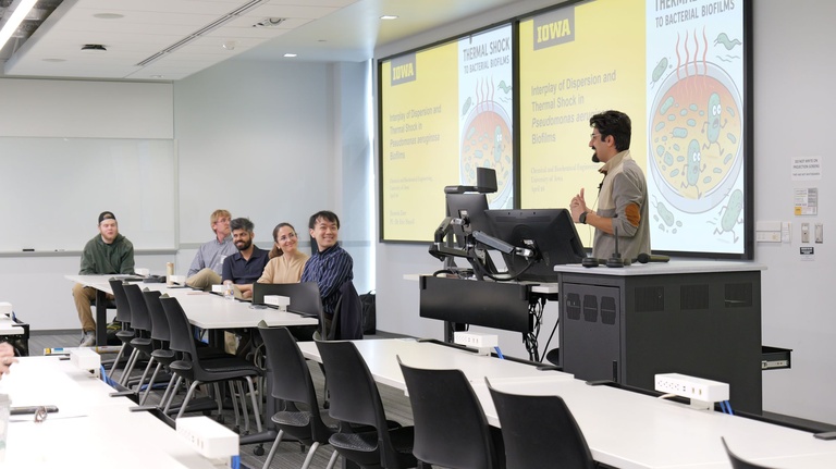 A presenter stands at a lectern in a classroom while several audience members sit at tables; presentation slides with University of Iowa branding and scientific diagrams are projected behind the speaker.