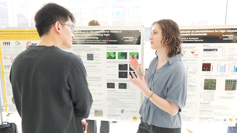 Two people discuss a research poster featuring images and charts, with one person gesturing while explaining the work in a sunlit academic space.