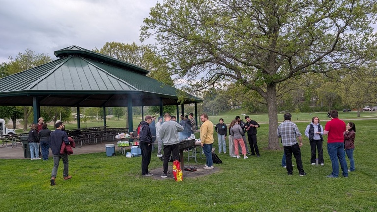 People stand and talk around a grill near a park pavilion during an outdoor gathering, with picnic tables, trees and open green space in the background.