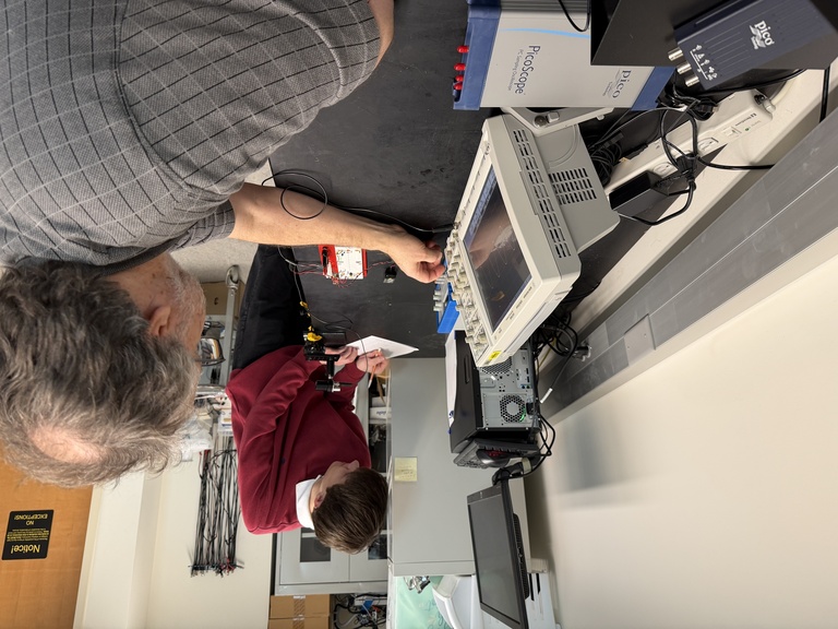 Older man, seated, twists a dial on a monitor while younger man wearing red sweater takes notes