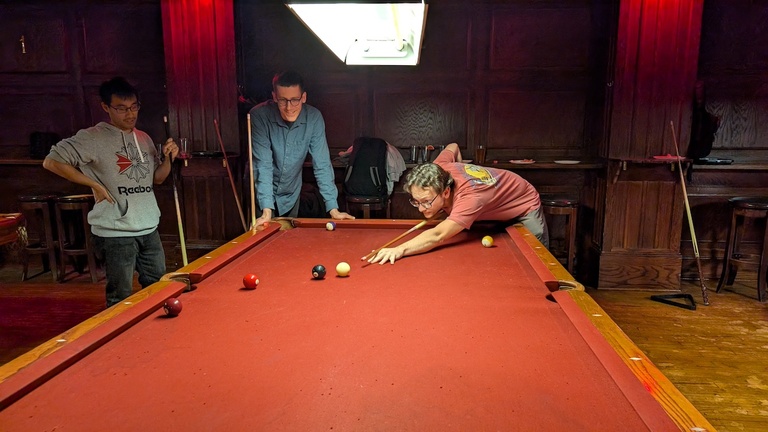 Several people gathered around a pool table as one player takes a shot in a dimly lit bar.