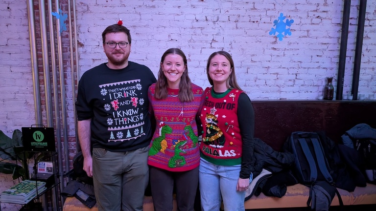 Three people posing against a brick wall decorated with snowflakes, wearing colorful holiday sweaters.
