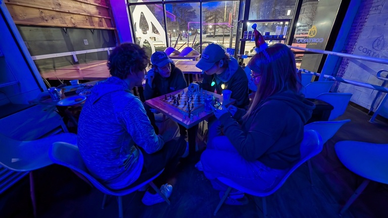 Four people seated at a café table playing chess under blue lighting, with snowy streets visible outside.