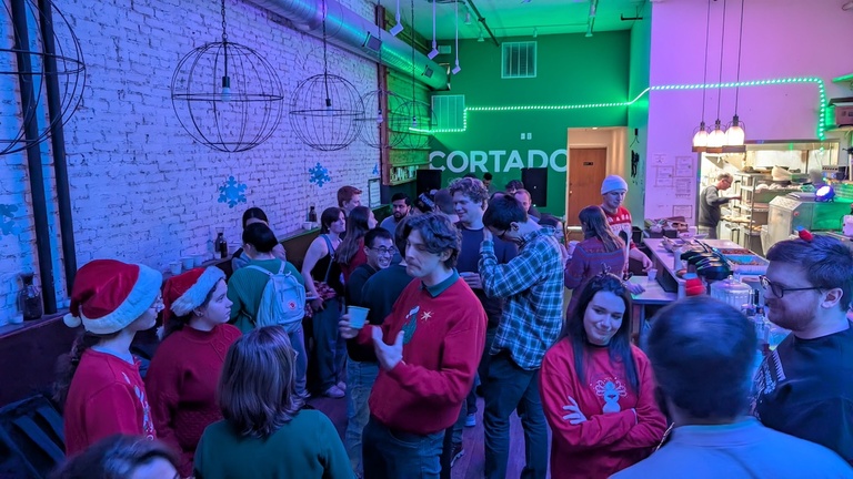 Holiday party crowd gathered inside a café with colorful lighting, exposed brick walls, and festive sweaters.