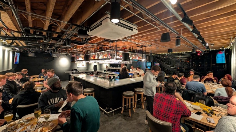 Wide view of a busy trivia night with people seated at tables and around a central bar in a modern, industrial-style space.