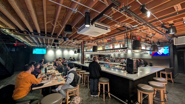 People gathered along a long bar and at nearby tables during a trivia event, with overhead lighting and shelves of bottles behind the bar.