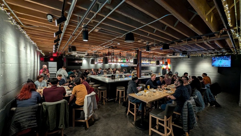 Crowded trivia night scene showing multiple tables filled with participants in a large room with exposed wood ceiling beams and string lights.