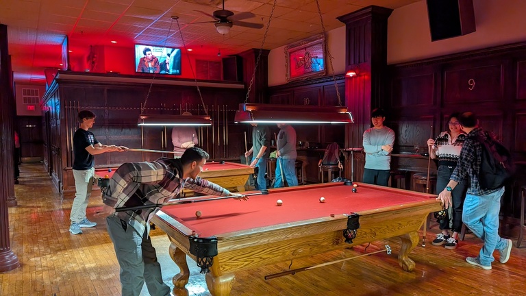 Wide view of a bar with two pool tables, showing multiple people playing and watching.