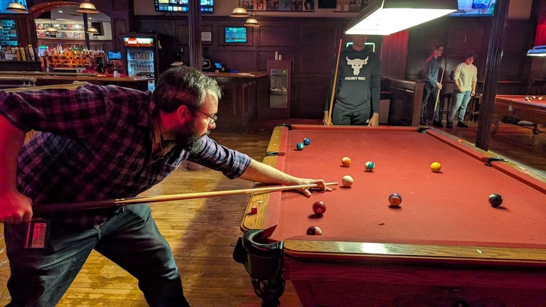 Person lining up a shot on a pool table inside a bar with dark wood walls and hanging lights.