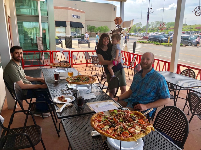 People gathered at an outdoor patio table with pizzas and drinks near a shopping center.