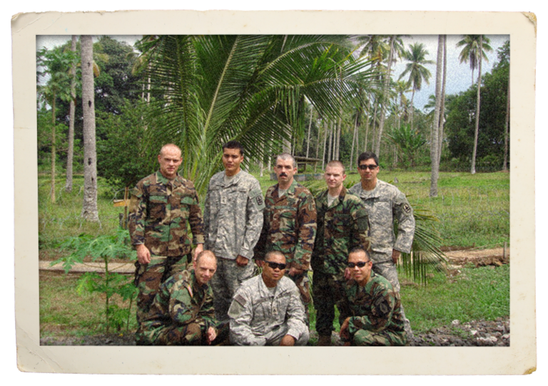 A group of eight men in army gear pose in front of palm trees