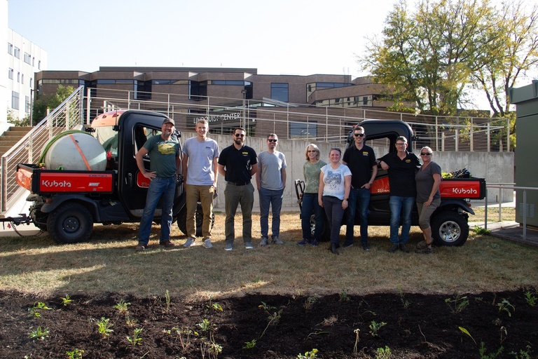Group of people between two utility vehicles beside a newly planted pollinator garden outside a campus building.