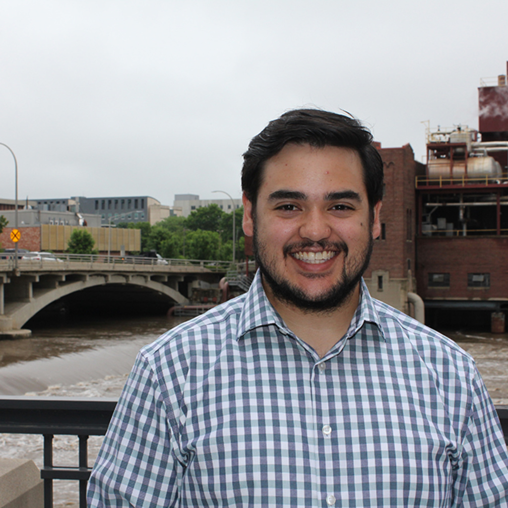 PhD student Isaac di Napoli smiles in front of the Iowa river in a checkered shirt