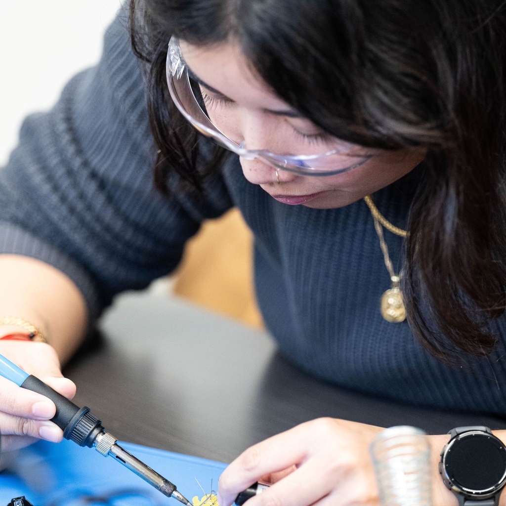 Student wearing safety glasses soldering components onto a small circuit board.