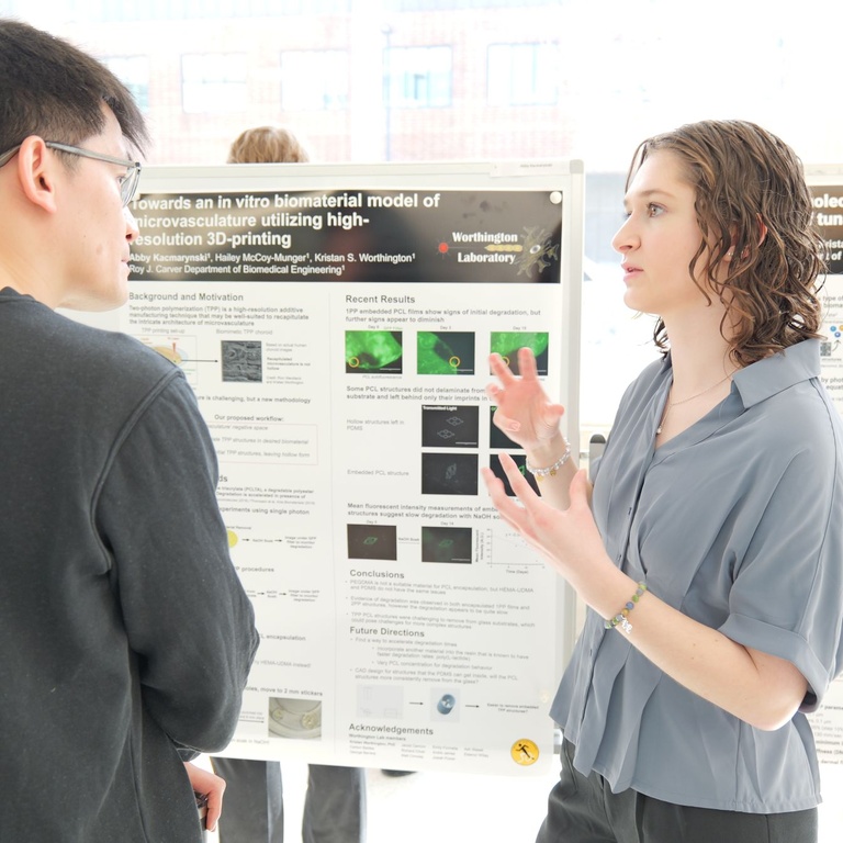 Two people discuss a research poster featuring images and charts, with one person gesturing while explaining the work in a sunlit academic space.