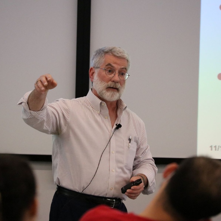 Speaker presenting at a lecture, gesturing toward a slide while holding a remote, with an audience in the foreground.
