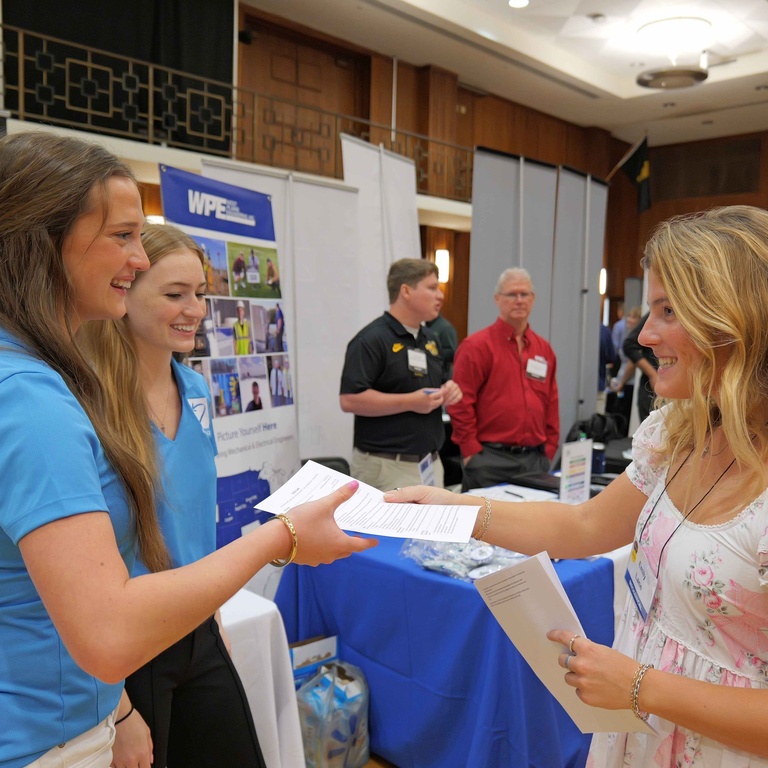 A University of Iowa student hands a resume to employer representatives at the Engineering Career Fair. The interaction takes place among recruiting booths in the Iowa Memorial Union ballroom.