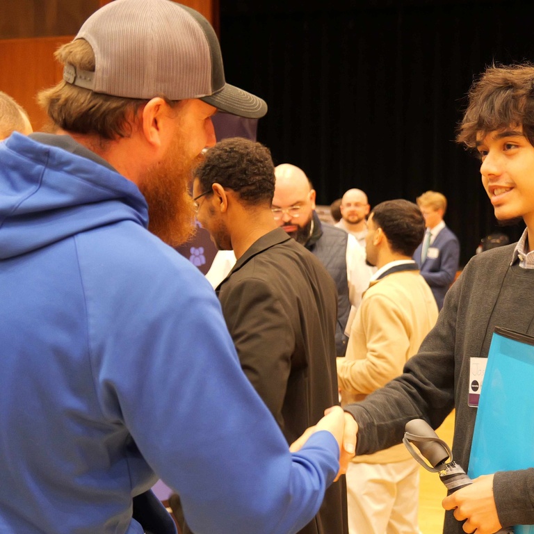 A University of Iowa engineering student holding a blue portfolio shakes hands with an employer representative at a booth during the Engineering Career Fair in the Iowa Memorial Union. Other students and recruiters talk in the background.