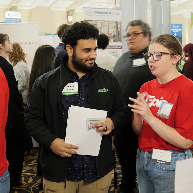 A University of Iowa engineering student holding a resume talks with representatives from Epic during the Engineering Career Fair. Additional students and employers network throughout the crowded event space.