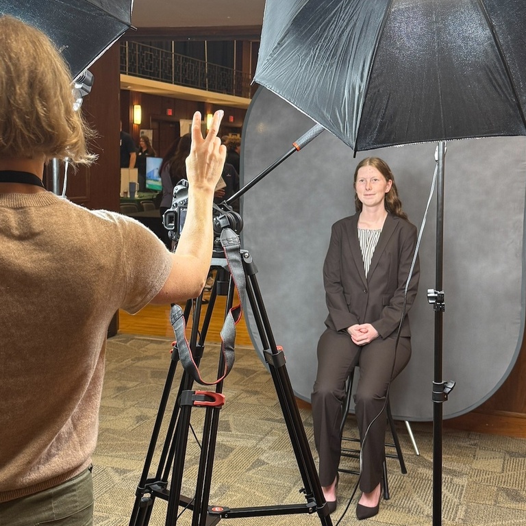 A University of Iowa student sits for a professional headshot during the Engineering Career Fair at the Iowa Memorial Union. A photographer stands behind a camera and lighting setup while the student poses in business attire in front of a gray backdrop.
