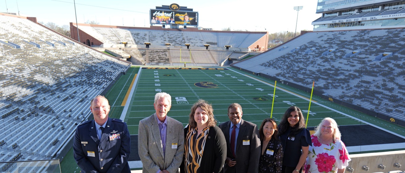 Seven people pose for a group photo in the seating area of a football stadium overlooking the field.