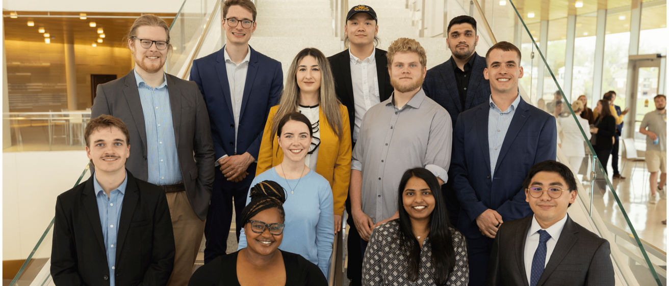 Group portrait of 12 professionally dressed individuals posed on a wide staircase inside a modern building with glass railings and a bright atrium. The group is arranged in three rows, with four people seated or standing in the front, four in the middle, and four in the back. The individuals wear business and business-casual attire in shades of blue, gray, black, and yellow. Large windows, warm wood accents, and several people in the distant background are visible.