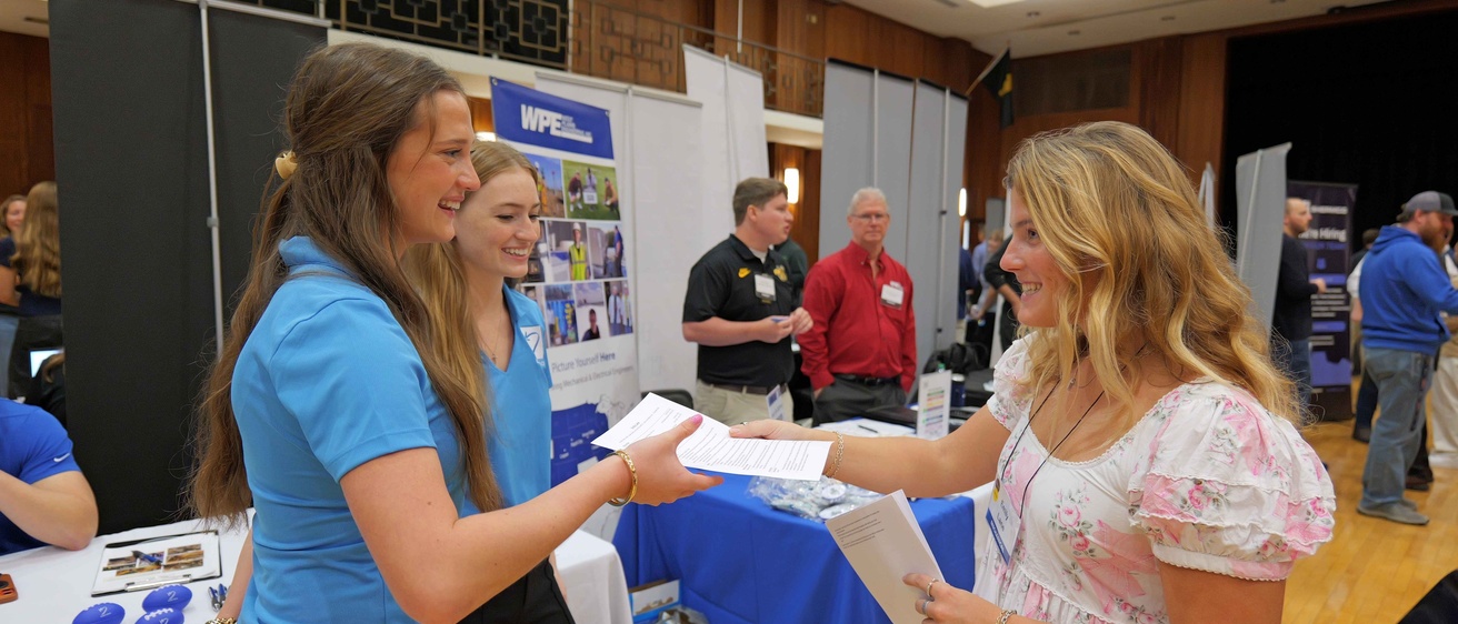A University of Iowa student hands a resume to employer representatives at the Engineering Career Fair. The interaction takes place among recruiting booths in the Iowa Memorial Union ballroom.