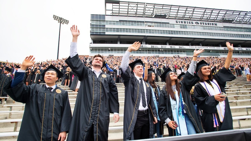 5 students wearing graduation caps and gowns standing on stadium bleachers waving their hands