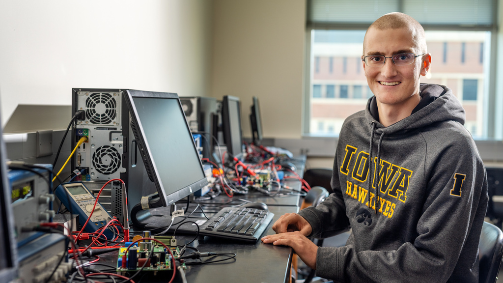 Noah Brown in grey and gold Iowa sweatshirt at a desk with computer monitor and a lot of wires