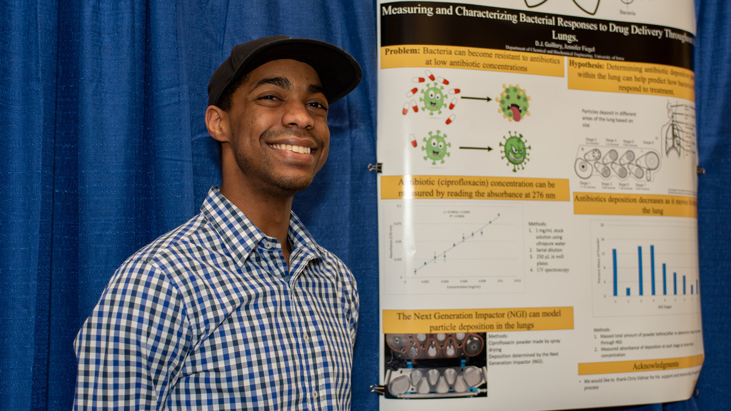 Man wearing hat and plaid shirt smiles next to research poster 