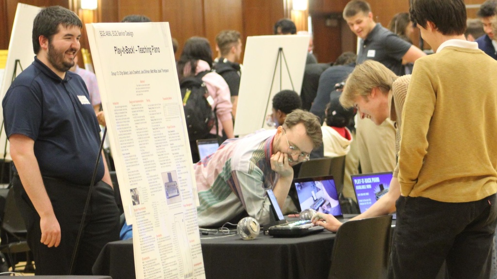 Students present a senior design project at a poster session, demonstrating software and hardware prototypes while discussing their work with attendees.