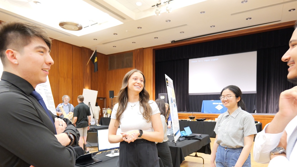 Group of people conversing at an indoor poster session with display boards and tables in a conference room.