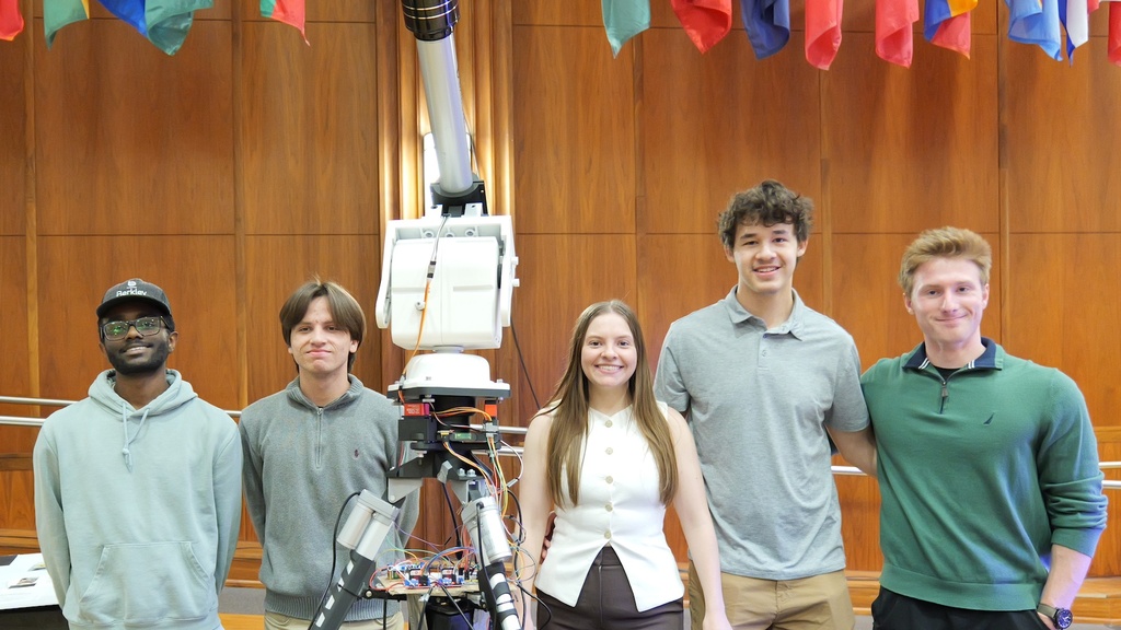 Five students stand beside a tall, tripod-mounted engineering device with exposed wiring and a cylindrical tube, in a wood-paneled room with international flags displayed overhead.