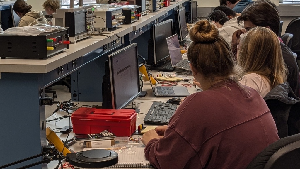 Students work at electronics lab stations with computers and testing equipment.