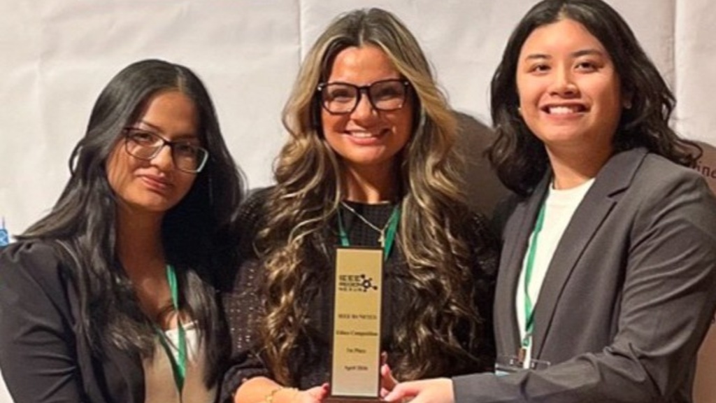 Three people in business attire hold an IEEE award plaque in front of a branded event backdrop.
