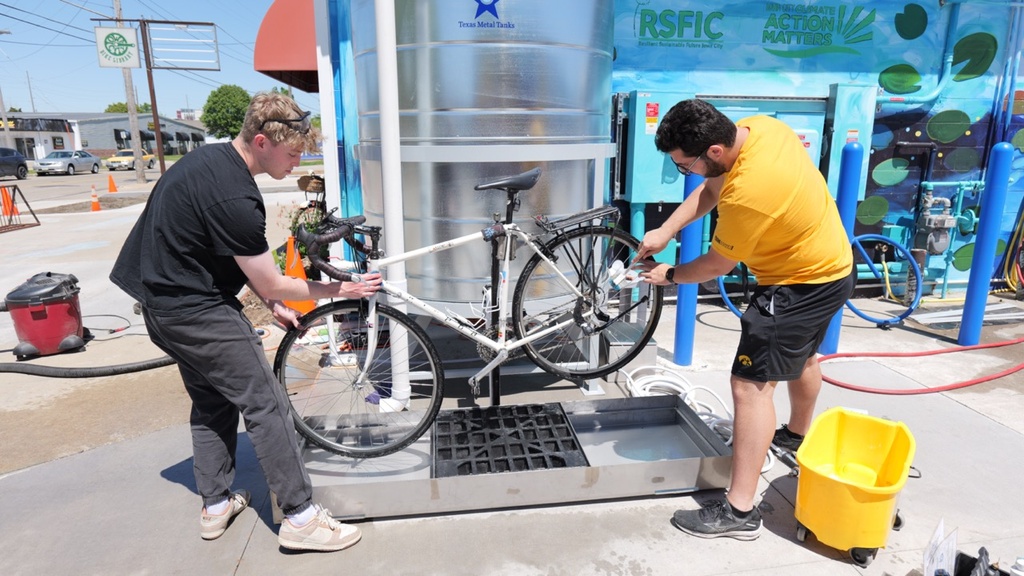 Man in yellow shirt and dark hair (right) and man in dark shirt and light hair (left) wash white bike in center with silver tank behind