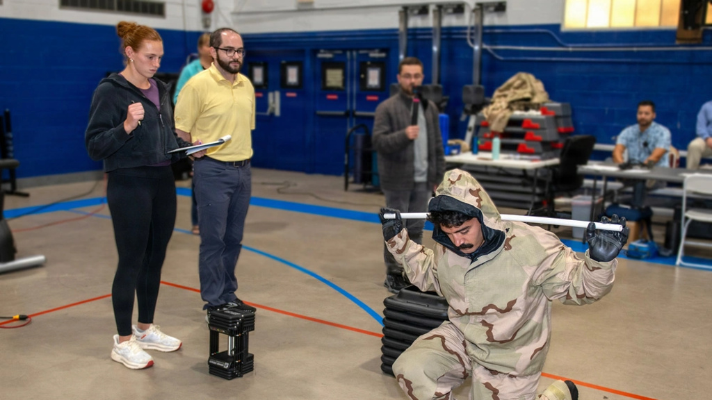 Research participant wearing full protective gear performs a kneeling squat with a bar while observers record data during a human performance study in an indoor lab setting.