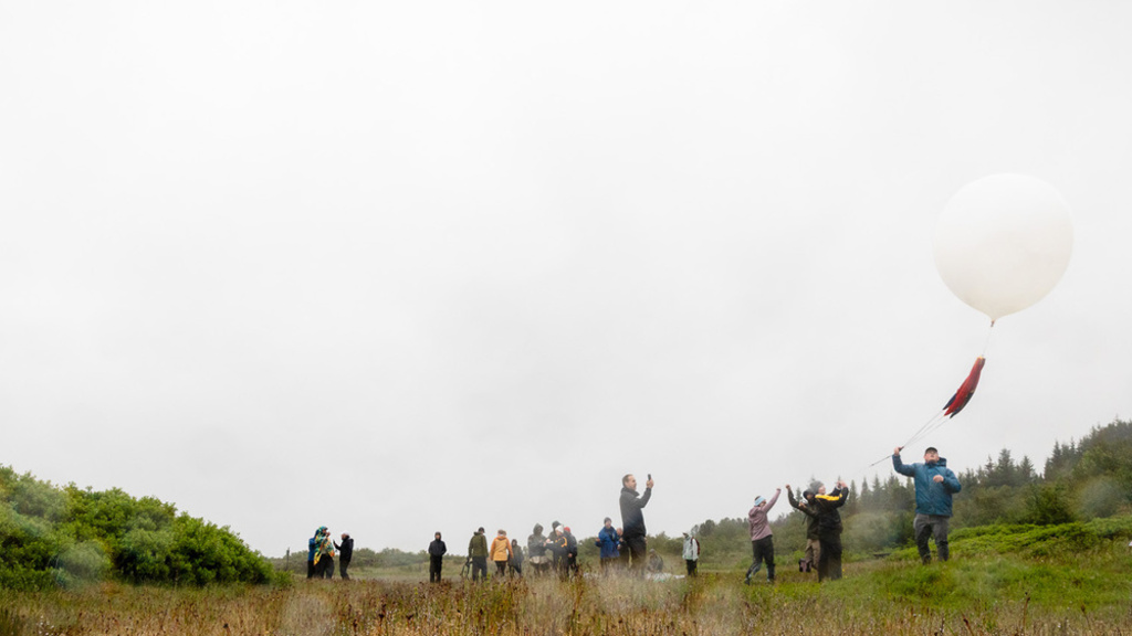 Group of people in a grassy field launching a large white weather balloon on a cloudy, rainy day.