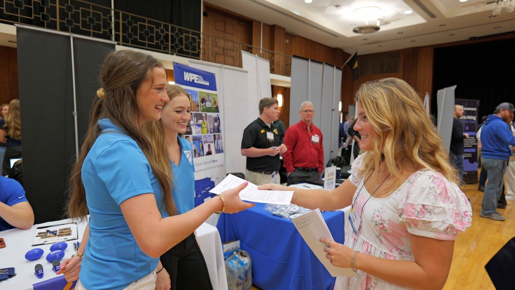 A University of Iowa student hands a resume to employer representatives at the Engineering Career Fair. The interaction takes place among recruiting booths in the Iowa Memorial Union ballroom.