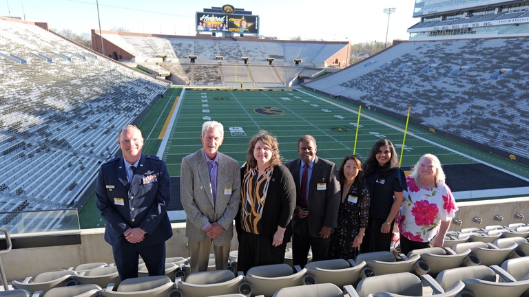 Seven people pose for a group photo in the seating area of a football stadium overlooking the field.