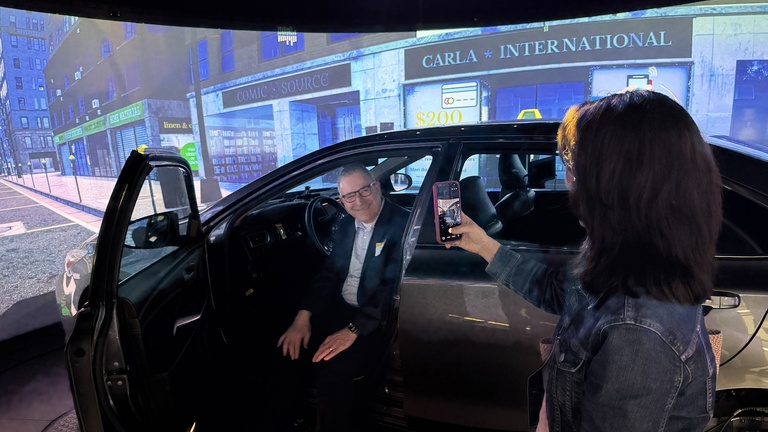 Person seated in a vehicle inside a driving simulator as another person takes a photo with a smartphone, surrounded by a projected city street scene.