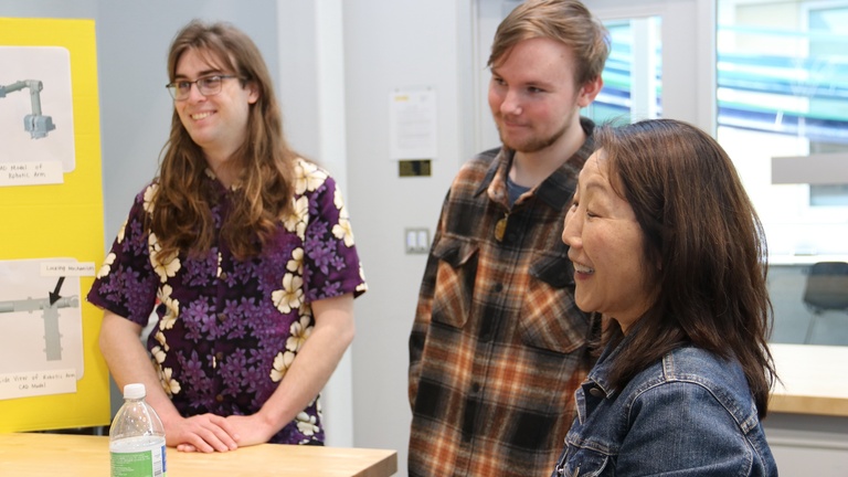 Two students present a robotics project to a visitor in a laboratory workspace.