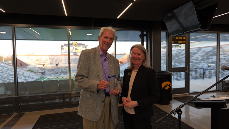 Two individuals pose with an award during a recognition event overlooking Kinnick Stadium.